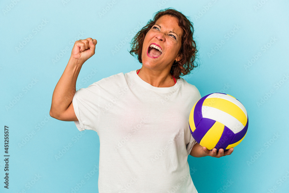 Middle age latin woman playing volleyball isolated on blue background raising fist after a victory, winner concept.