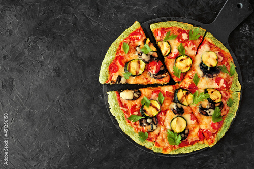 Healthy, low carb broccoli crust pizza with tomatoes, zucchini and mushrooms. Overhead view with cut slices on a dark slate background.