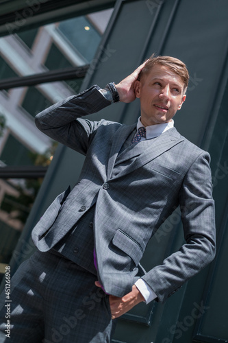 Portrait of handsome young businessman in grey classic suit. Standing near dark green building, touching his head and watching aside. © Aleksandr