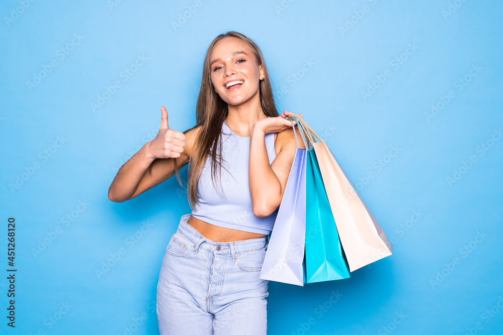 Young woman over isolated blue background holding shopping bags and with thumb up Young woman over isolated blue background holding shopping bags and with thumb up