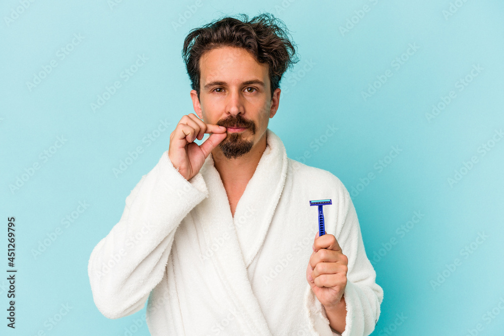 Young caucasian man holding a razor blade isolated on blue background with fingers on lips keeping a secret. Young caucasian man holding a razor blade isolated on blue background with fingers on lips keeping a secret.