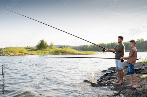 Teenage boy and his father fishing together by lake on summer weekend