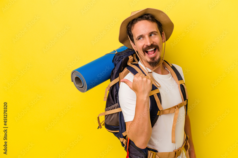 Young caucasian hiker man isolated on yellow background points with thumb finger away, laughing and carefree.