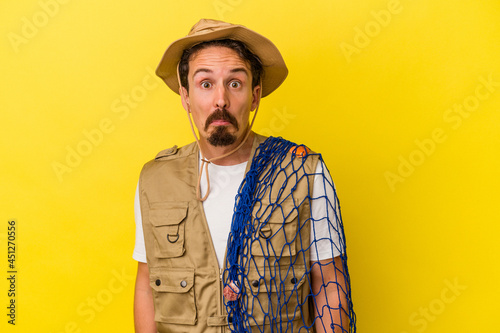 Young caucasian fisherman holding net isolated on yellow background shrugs shoulders and open eyes confused.