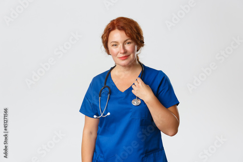 Smiling caring middle-aged redhead doctor, female physician in blue scrubs with stethoscope smiling at camera, treating patients in hospital, testing people for covid-19, standing grey background photo