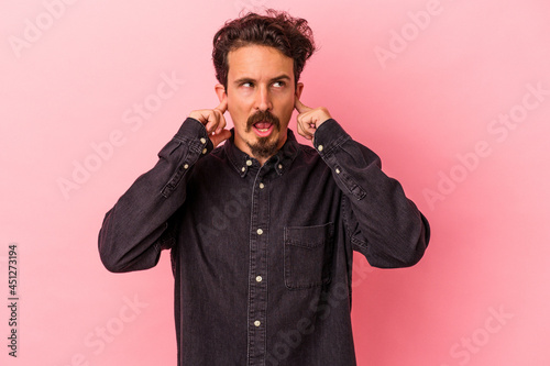 Young caucasian man isolated on pink background covering ears with fingers, stressed and desperate by a loudly ambient. © Asier