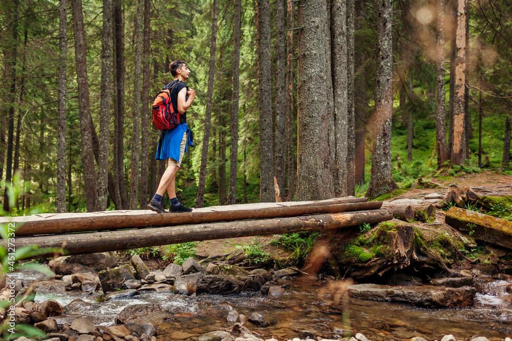 Fototapeta premium Traveler hiker with backpack crosses mountain river in Carpathian forest walking on bridge. Trip to summer Ukraine