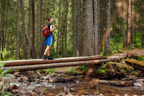Traveler hiker with backpack crosses mountain river in Carpathian forest walking on bridge. Trip to summer Ukraine