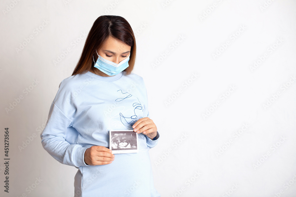 A pregnant brunette in a medical mask holds a picture of the baby's ultrasound in her hands. White background, space for text. Pregnancy monitoring. Screening. A pregnant brunette in a medical mask holds a picture of the baby's ultrasound in her hands. White background, space for text. Pregnancy monitoring. Screening.