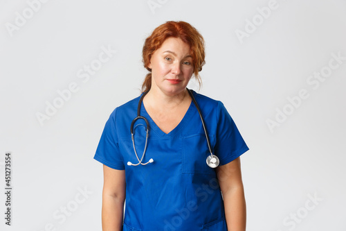 Medicine, healthcare and coronavirus concept. Skeptical and unamused redhead female medical worker, doctor smirk and looking reluctant with dismay, standing in blue scrubs over grey background photo