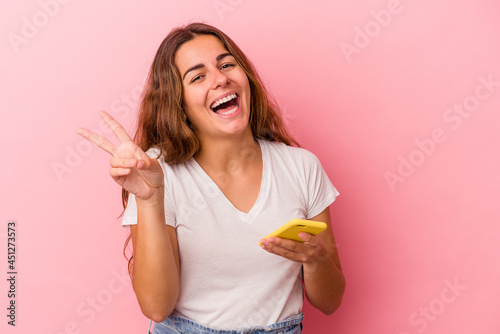 Young caucasian woman using mobile phone isolated on pink background joyful and carefree showing a peace symbol with fingers.