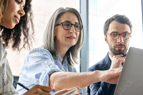Mature older ceo businesswoman mentor in glasses negotiating growth business plan with diverse executive managers at boardroom meeting table using laptop. Multicultural team work together in office. photo