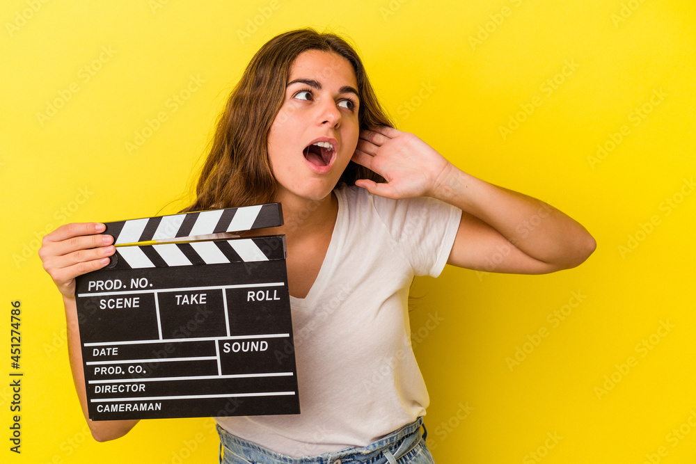 Young caucasian woman holding clapperboard isolated on yellow background  trying to listening a gossip.