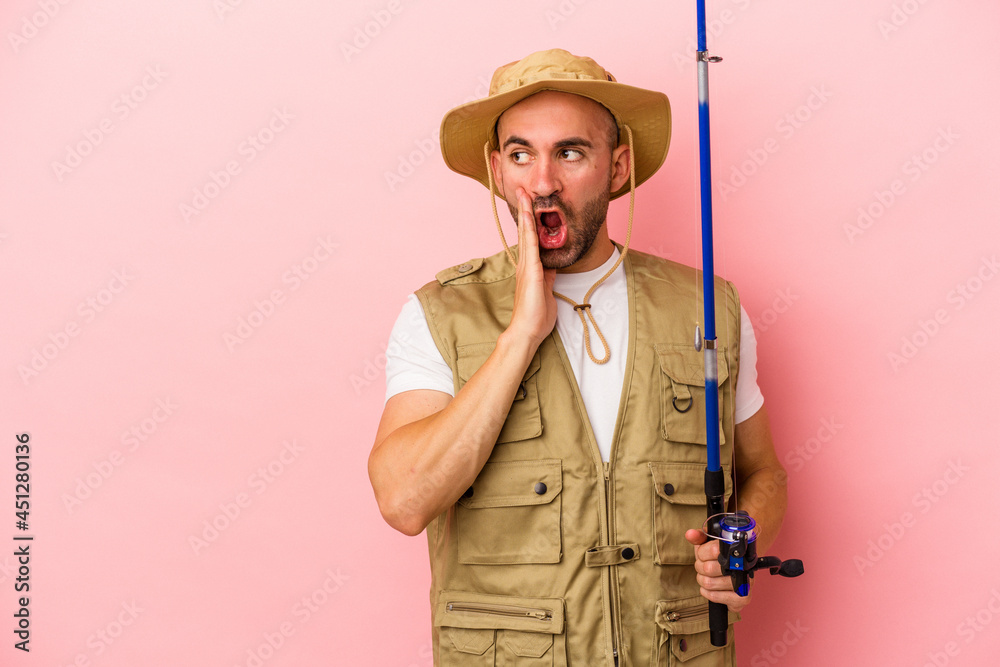 Young bald fisherman holding a rod isolated on pink background is saying a secret hot braking news and looking aside Young bald fisherman holding a rod isolated on pink background is saying a secret hot braking news and looking aside