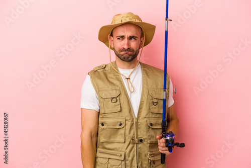 Young bald fisherman holding a rod isolated on pink background confused, feels doubtful and unsure.
