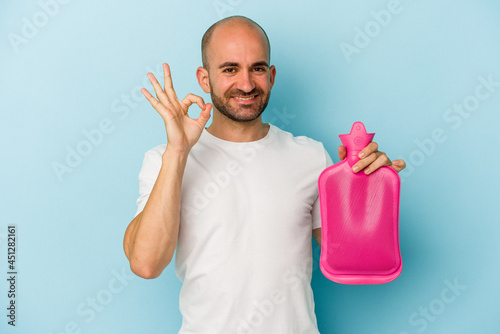 Young bald man holding a hot bag of water isolated on blue background cheerful and confident showing ok gesture.