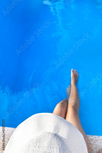 Top view of a woman wearing a white hat seated on the edge of a swimming pool. She gently plays with the water moving her legs. Summer relax concept.