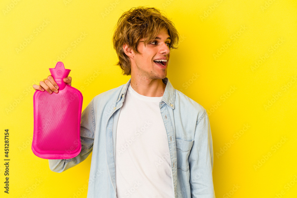 Young caucasian man with makeup holding a hot bag of water isolated on yellow background looks aside smiling, cheerful and pleasant. Young caucasian man with makeup holding a hot bag of water isolated on yellow background looks aside smiling, cheerful and pleasant.