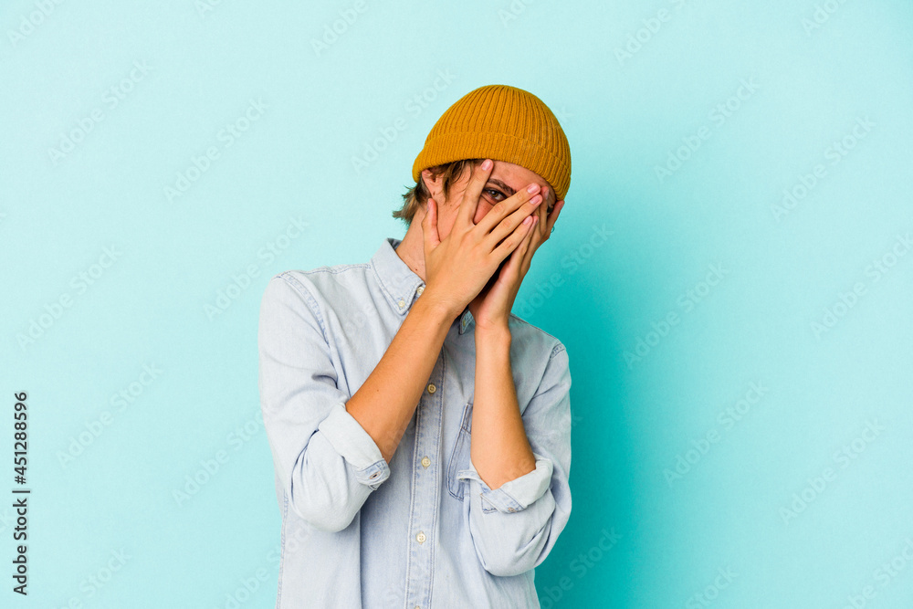 Young caucasian man with make up isolated on blue background blink at the camera through fingers, embarrassed covering face. Young caucasian man with make up isolated on blue background blink at the camera through fingers, embarrassed covering face.