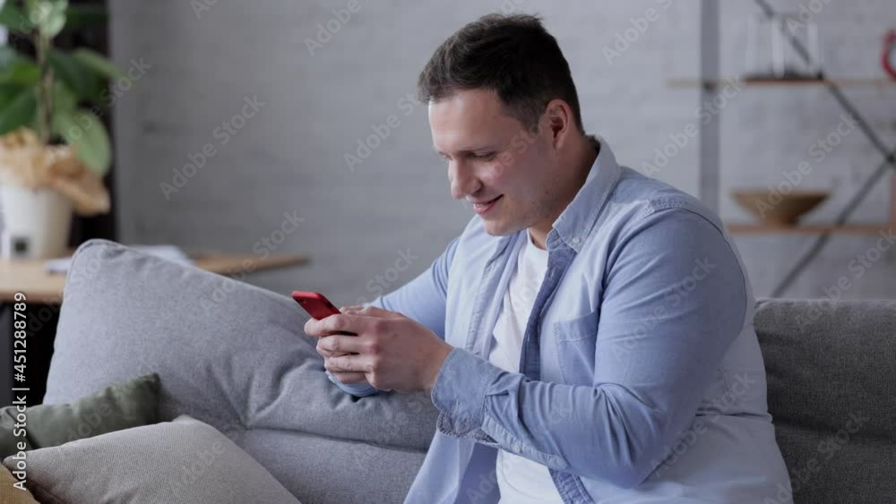 Smiling Male millennial holding modern smartphone texting message. Happy Young Man Uses Smartphone while Sitting on a Sofa at Home. Cheerful Caucasian guy texting message and typing on the smartphone.