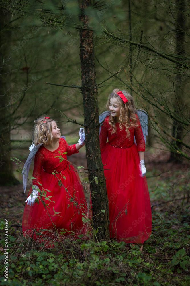 Zwei Engel schauen sich aus dem Baum an und lachen. Sie spielen und freuen sich. Sie haben rote Kleider an, weiße Flügel und rote Schleife in der Haaren, blonde Locken und weiße Handschuhe. 