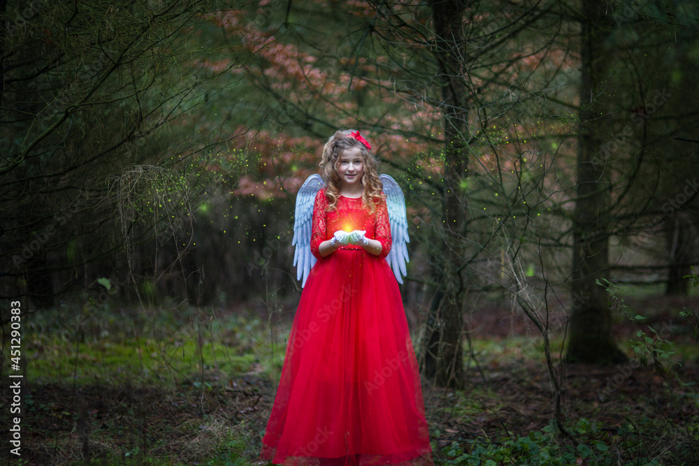 Weihnachtsengel in einem Märchen Wald. Es liegt kein Schnee. Typisch für Europa. Sie hat Zauberlicht in der Hand. Das Engel hat rotes Kleid an, weiße Flügel und rote Schleife in der Hand