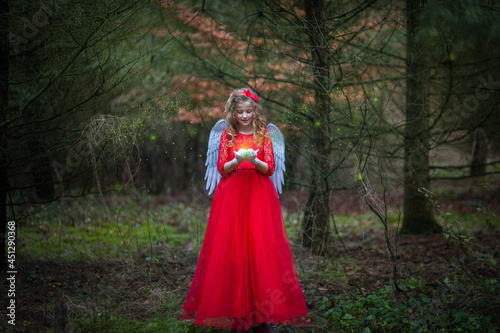  Weihnachtsengel in einem Märchen Wald. Es liegt kein Schnee. Typisch für Europa. Sie hat Zauberlicht in der Hand. Das Engel hat rotes Kleid, weiße Flügel und rote Schleife in der Hand. S 
