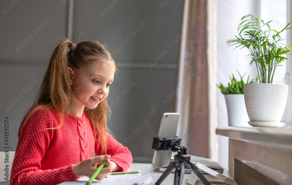 Little girl of european appearance looks at the smartphone screen at home with smile. Kids online, technology, online education, home studying and communication concept Little girl of european appearance looks at the smartphone screen at home with smile. Kids online, technology, online education, home studying and communication concept