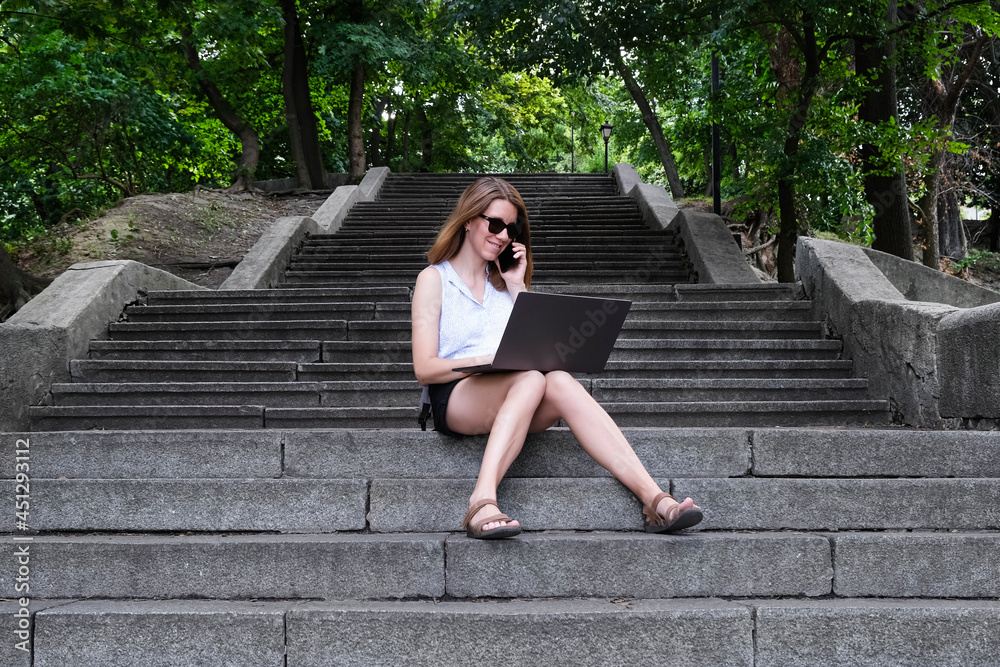 Young girl or woman sitting on the steps in the park and working on a laptop. Freelance, education or working online concept.