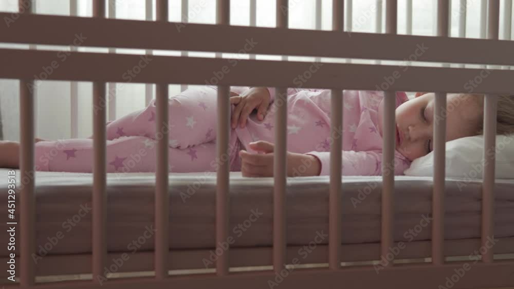 close up authentic cute caucasian little preschool child girl sleep sweetly in white crib on pajama in bedroom during lunchtime. kid is rest lie in bed near window. Childhood, medicine, health concept
