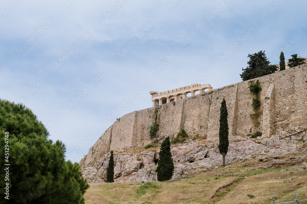 View from below of the Acropolis of Athens. Ancient Greek settlement. Greek mythology. Landmark of Athens. Green trees near high stone wall.