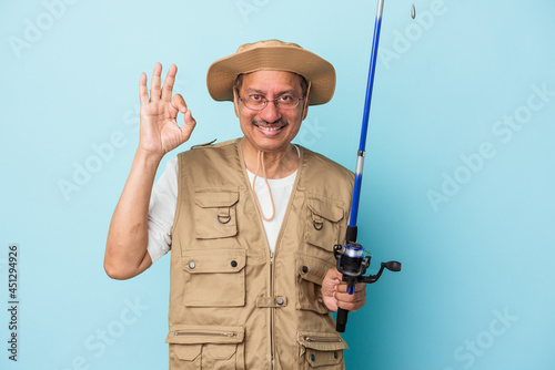 Senior indian fisherman holding rod isolated on blue background cheerful and confident showing ok gesture.