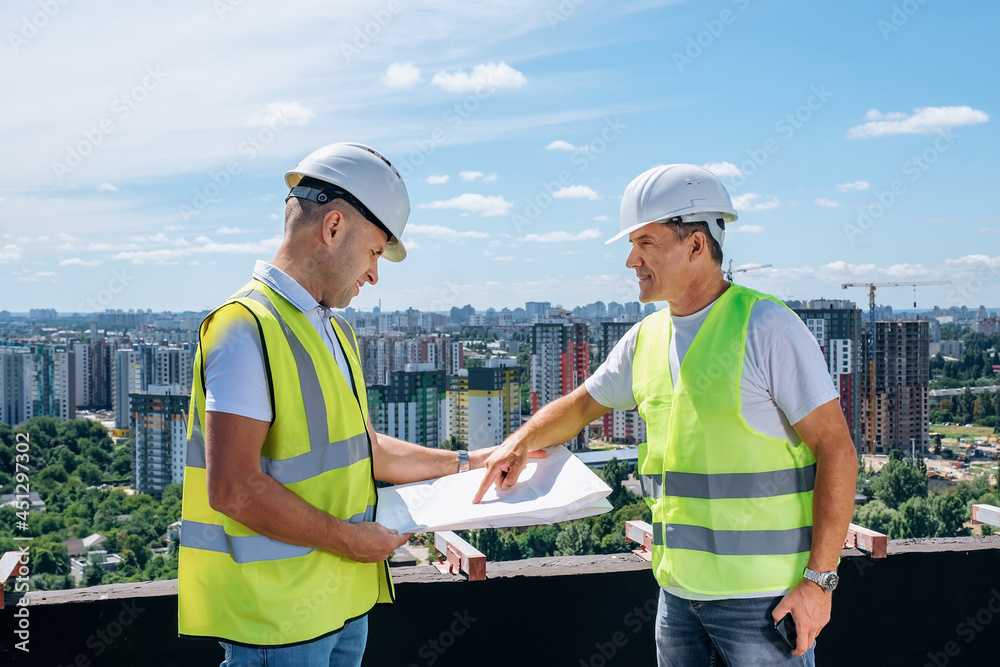 Adult men in uniform smiling and discussing draft while working on construction site on sunny day together Adult men in uniform smiling and discussing draft while working on construction site on sunny day together