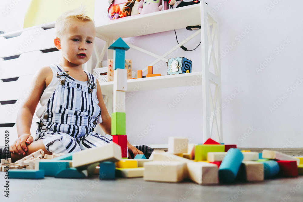 A boy with blonde hair in a jumpsuit plays children's educational wooden games in a bright room selective focus. A boy with blonde hair in a jumpsuit plays children's educational wooden games in a bright room selective focus.