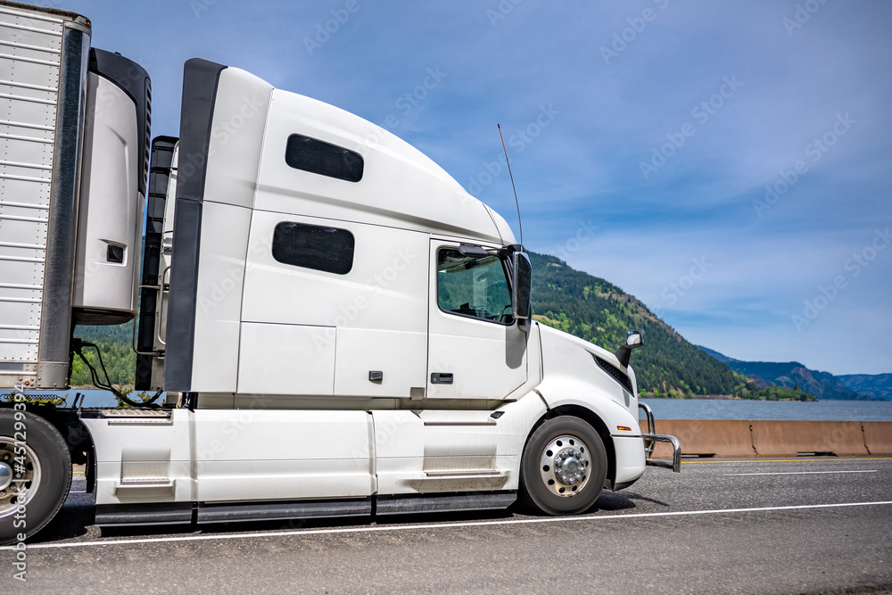 Profile of professional white big rig semi truck with reefer semi trailer with refrigerator unit on the front wall running on the road along the river
