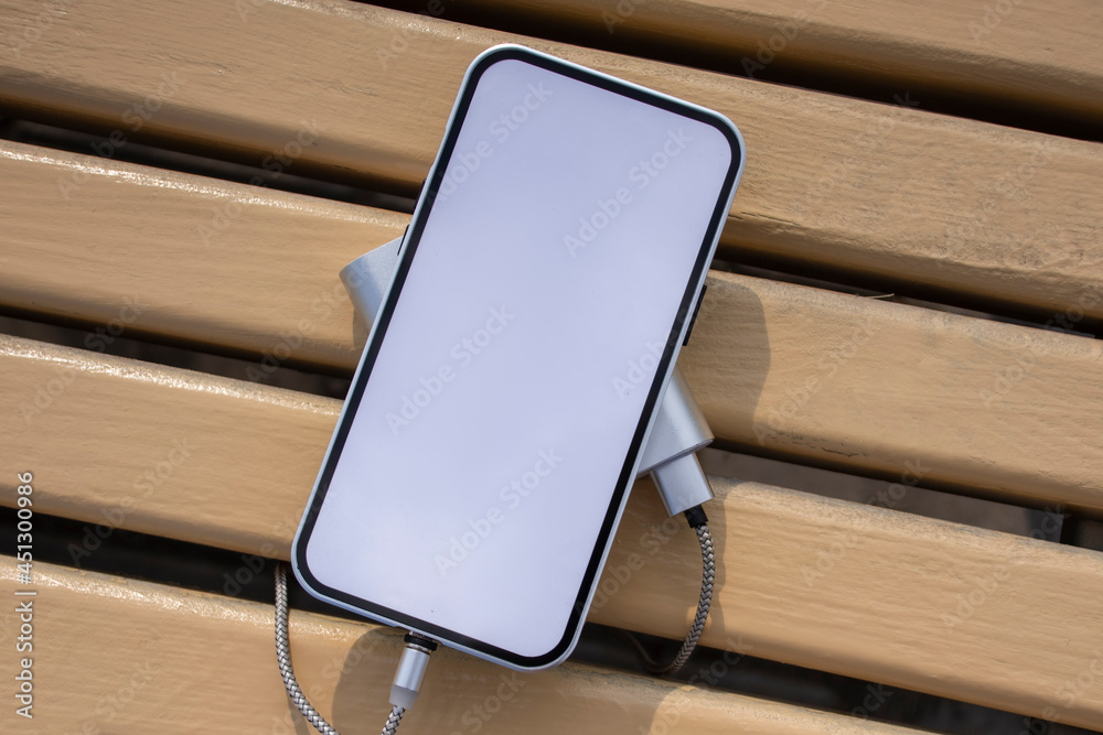 Power Bank charges a mockup smartphone with white screen on a wooden bench in the park. Power Bank charges a mockup smartphone with white screen on a wooden bench in the park.