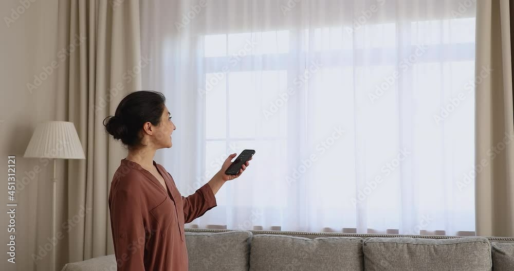 Young Indian woman close motorized window roller blinds in living room, holds cellphone use smart home automation mobile app, easy to control, contemporary house happy owner, modern tech usage concept