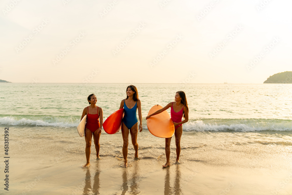 Confidence Asian woman girl friends in swimwear holding surfboard and walking together on the beach at summer sunset. Female friendship enjoy outdoor activity lifestyle play extreme sports surfing