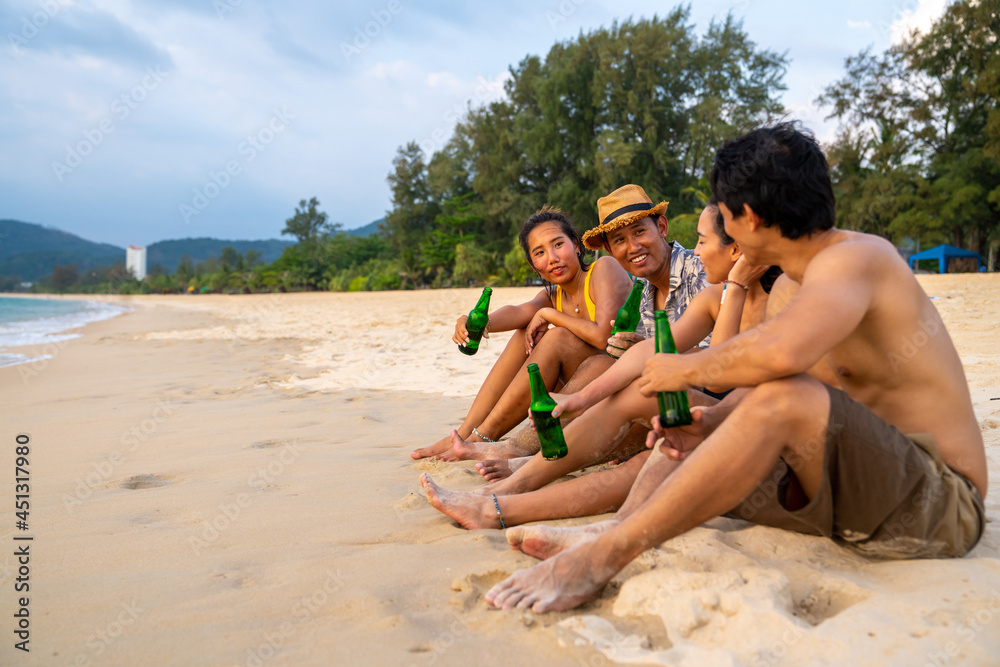 Fototapeta premium Group of Happy Asian man and woman friends sitting on the beach enjoy drinking beer with talking together at summer sunset. Male and female friendship relax and having fun outdoor lifestyle activity.