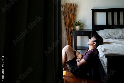 Depressed Asian man sit on the floor by window with stressed and hopeless face. Loneliness sad man living alone in bedroom with negative emotion. Mental health, economic and life problem concept