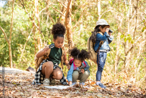 Group of Diversity little girl friends with backpack hiking together at forest mountain in summer sunny day. Three kids having fun outdoor activity sitting and looking at the map exploring the forest.