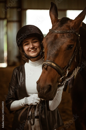 Young woman rider with a brown beautiful horse, standing nearby in jockey clothes, selective focus. A beautiful young girl stands next to a horse in the stable. She is wearing a beautiful white dress.