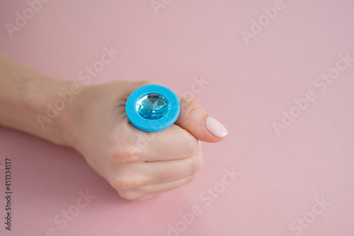 A woman is holding a blue anal plug with a crystal on a pink background. Adult toy for alternative sex