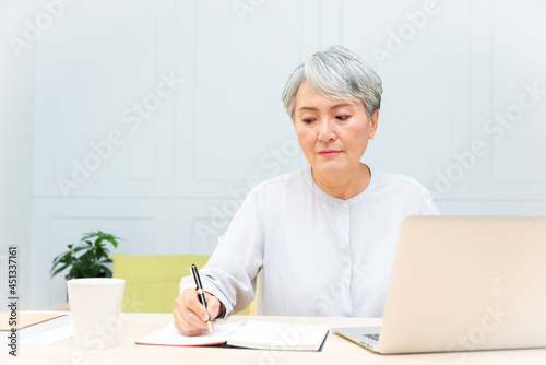 Senior asian woman at phone works on her laptop at home.