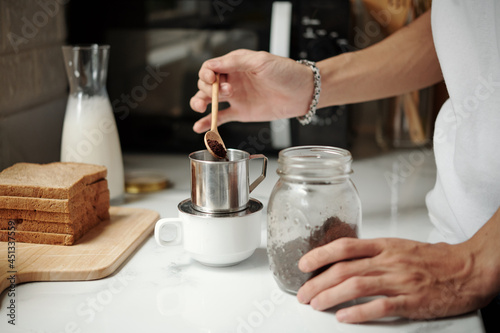 Hands of young man putting spoon of ground coffee in aluminum dripper on kitchen table
