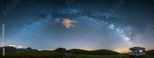 Panoramic night sky over Campo Imperatore highlands, Abruzzo, Italy. The Milky Way galaxy arc and stars over illuminated camper van. Camping freedom in unique hills landscape. photo