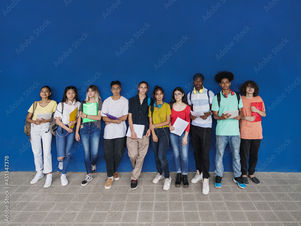 Obraz premium Group of multiracial teenager students looking to camera standing over blue wall copy space. Teenage multi ethnic classmates friends diversity in education