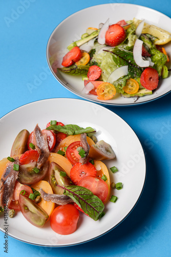 Two plates of vegetable salad made of tomatoes and fish slices, served in white plates over blue background.