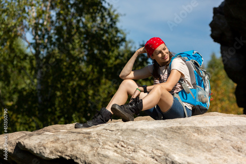 Hiker takes a rest. Caucasian tired woman with a backpack and red bandana sitting on a rock and smiling in sunny day in summer, selected focus.