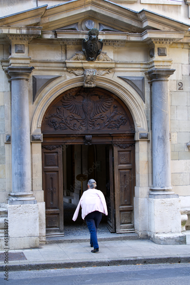 Elderly woman entering wooden door at medieval town hall building at City of Geneva on a sunny summer morning. Photo taken July 29th, 2021, Geneva, Switzerland.